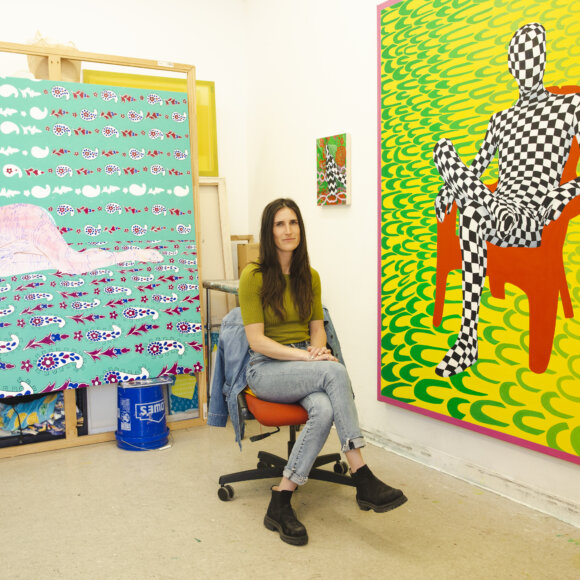 Artist, Taylor Chapin, seated in her studio next to her work, figures seated or lying down on colorful and patterned backgrounds.