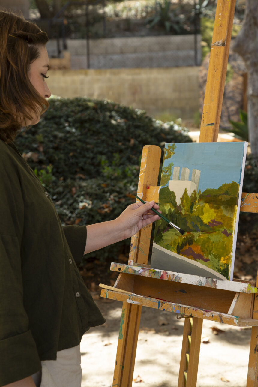 The image shows an artist standing outdoors, holding a paintbrush and working on a canvas mounted on an easel. The artist is dressed in a dark jacket and has a focused expression, seemingly absorbed in their work. The palette in their left hand contains several bright colors, including shades of green, yellow, and brown. In the background, the setting appears to be a lush, green park or garden, with soft, blurred foliage and a clear blue sky. The light is bright and natural, highlighting the artist and their canvas. The brushstrokes on the canvas suggest the artist is capturing the colors and textures of the landscape before them.